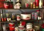 Three shelves with strategically displayed items one could find at the vintage market all following a red and white colour scheme, items such as: metal apples, a panda bear, a bottle, thermoses, owls, salt and pepper shakers, a Campbell’s soup mug, some pots and bowls, and a plate with a corgi and a kitten.