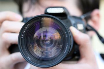 This is a close-up photo of a person holding a camera. The camera is black and the lens is front and center. A person holds the camera to their face, but the person is blurred out. 